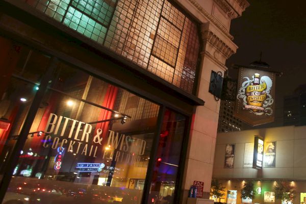 A street view at night shows the facade of "Bitter & Twisted Cocktail Parlour" with lit signage and reflections on the glass windows, creating a vibrant atmosphere.