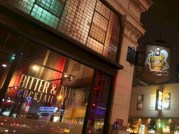 A street view at night shows the facade of "Bitter & Twisted Cocktail Parlour" with lit signage and reflections on the glass windows, creating a vibrant atmosphere.