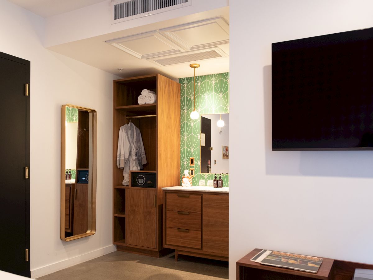 This image shows a modern hotel room with a wall-mounted TV, a mirror, and a wardrobe area with a safe and a hanging robe.