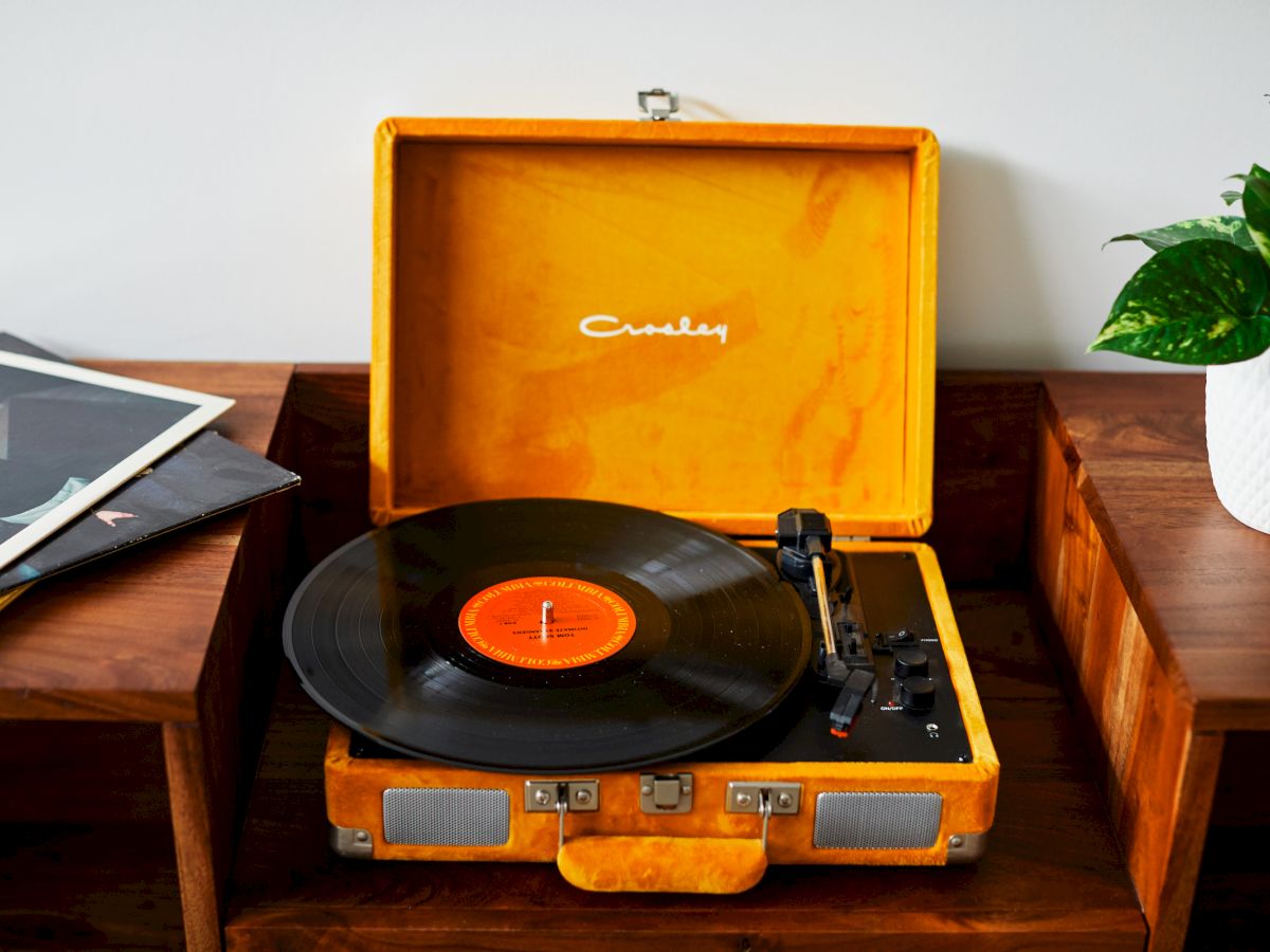 A vintage record player with a vinyl record is placed on a wooden table next to a stack of albums and a potted plant.