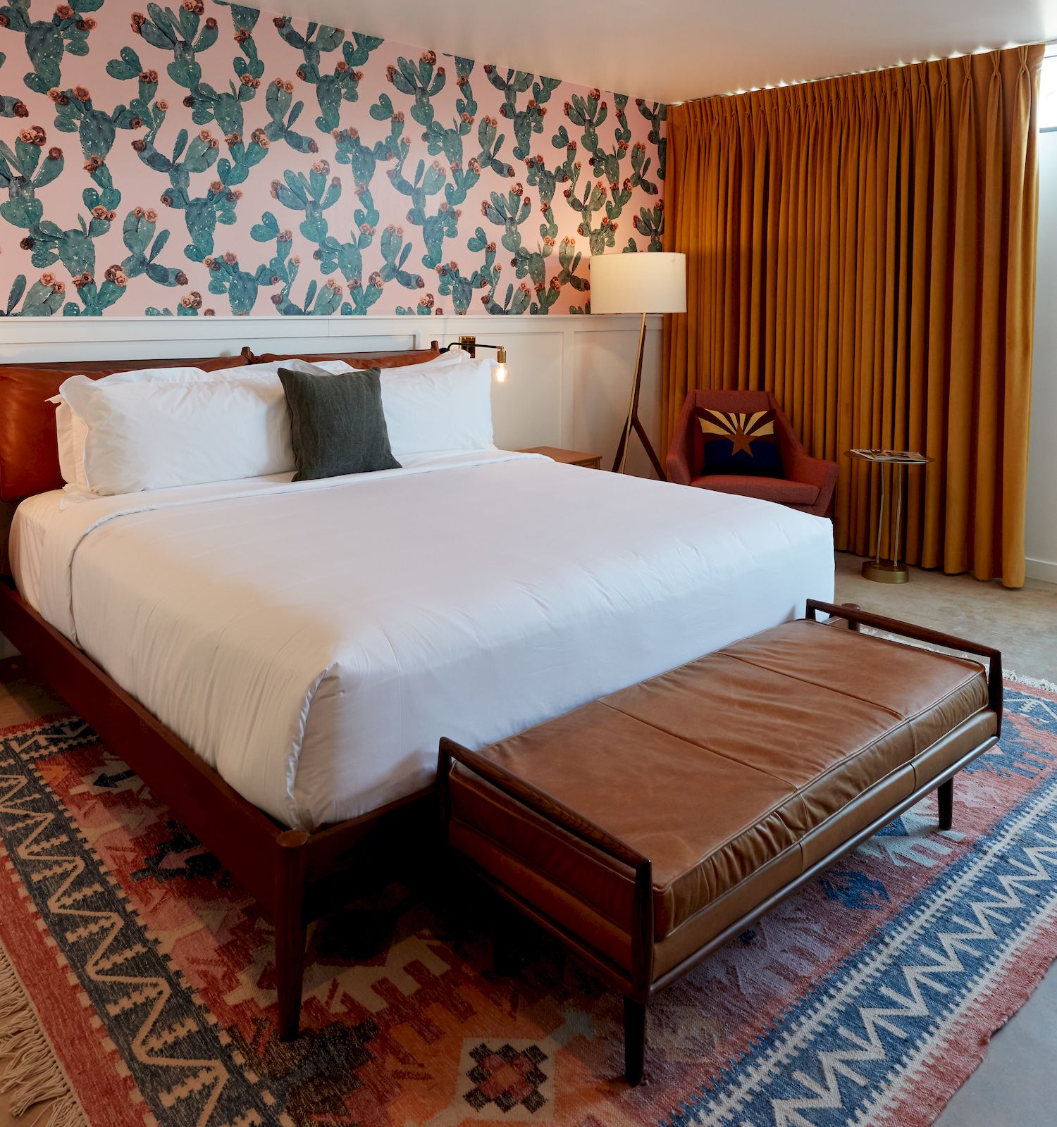 A modern bedroom with a large bed, cactus-themed wallpaper, wooden furniture, a geometric rug, mustard curtains, and a sitting area with a lamp.