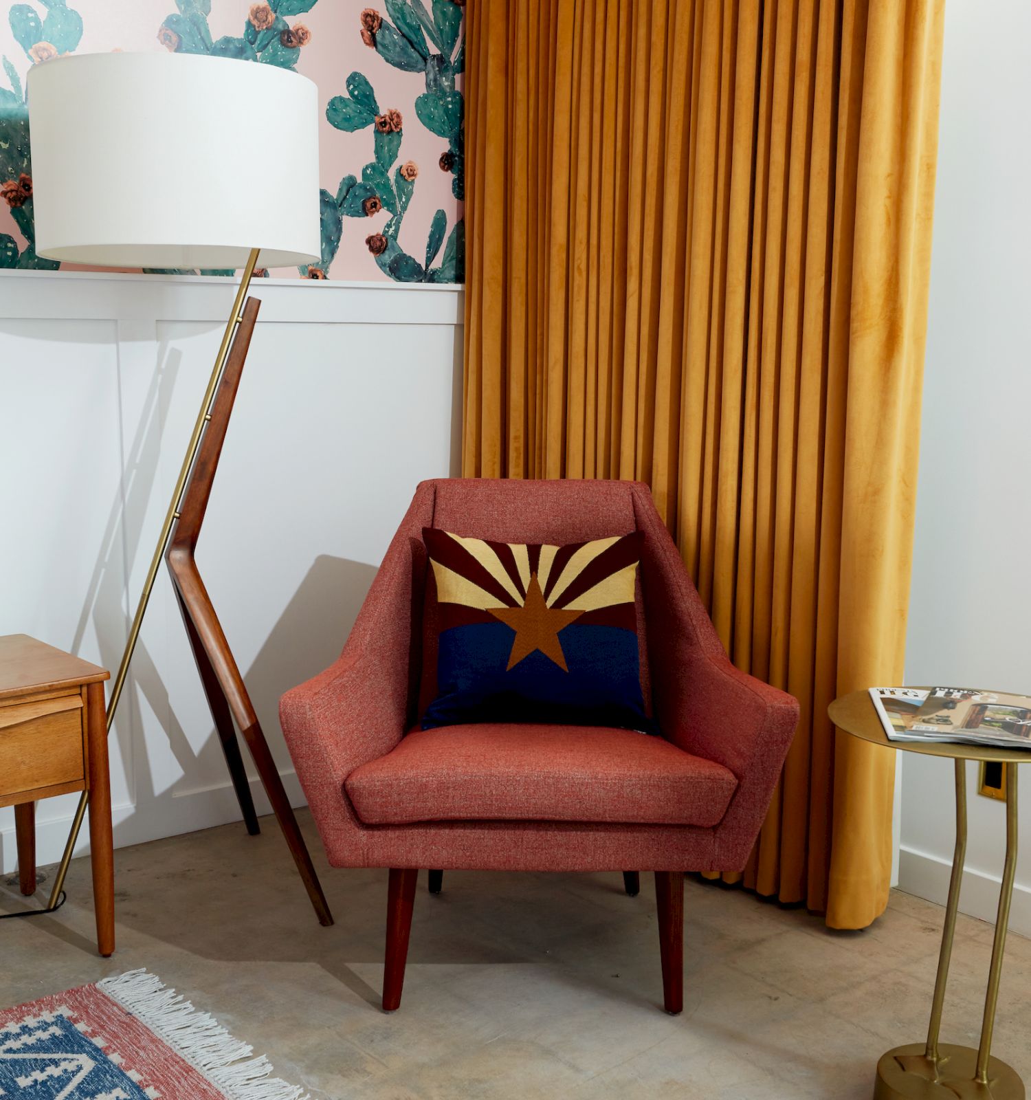 A cozy corner with an orange chair, decorative pillow, yellow curtains, floor lamp, cactus-themed wallpaper, and a small round table.