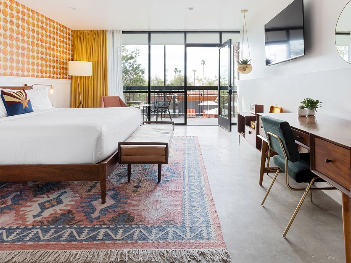 A modern hotel room with a large bed, retro wallpaper, a desk, a TV, and a balcony view. The room features stylish decor and a patterned rug.