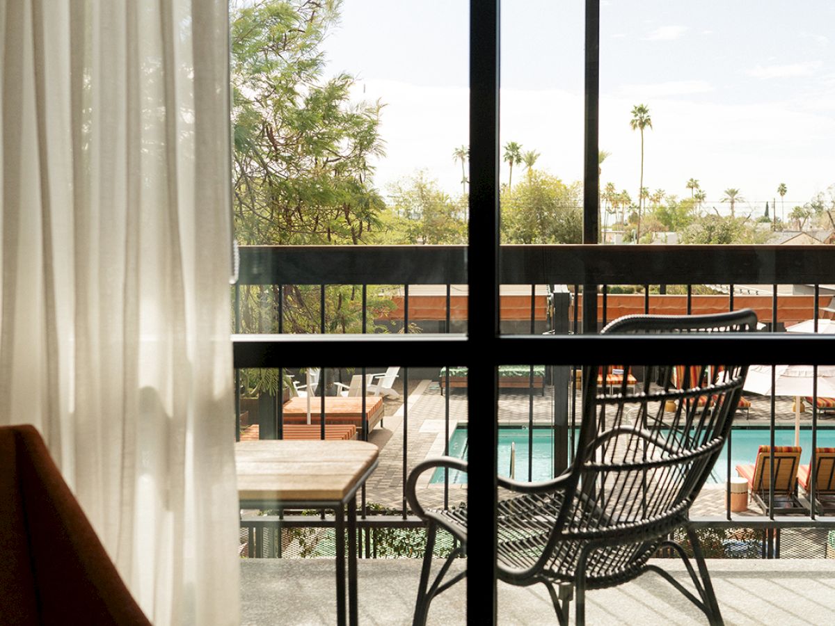 A balcony view with a black wire chair, looking out to a pool area. There are trees and palm trees visible in the background, with sheer curtains partially drawn.