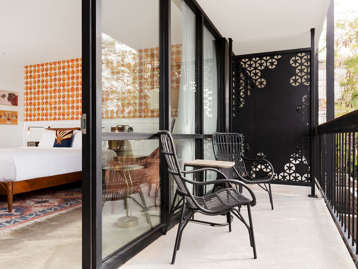 This image shows a modern room with a bed, an attached balcony with two black chairs, a small table, and vibrant orange patterned wallpaper ending the sentence.