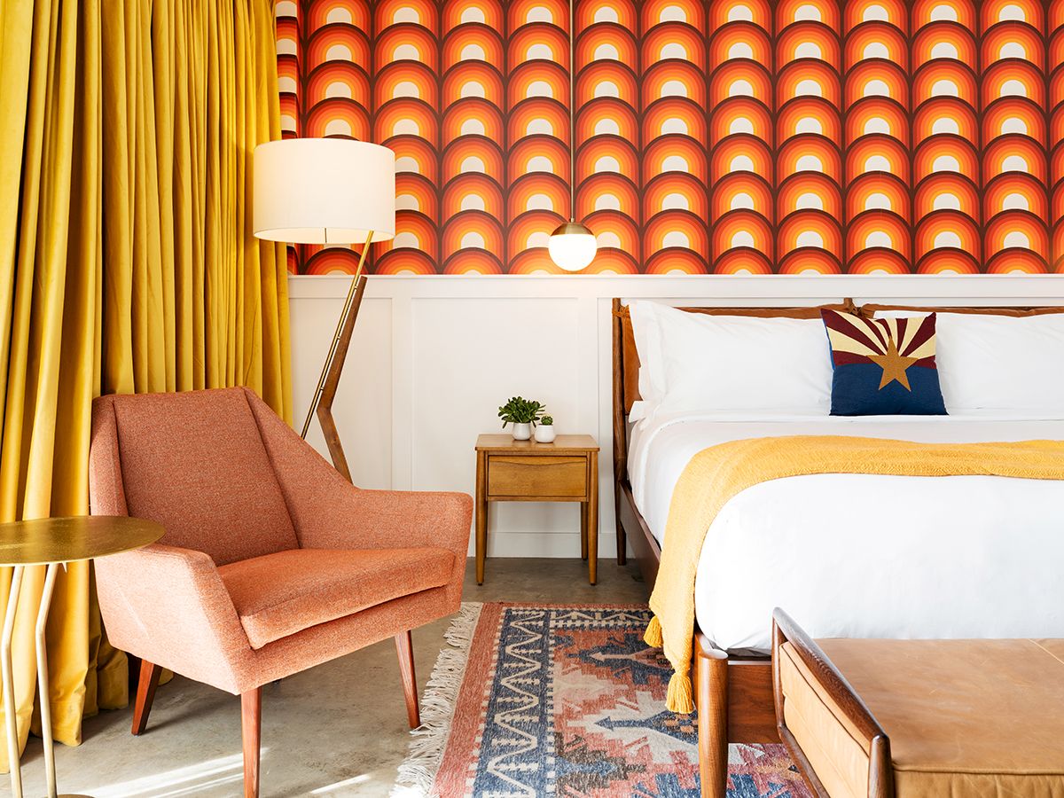 A mid-century modern bedroom with patterned wallpaper, mustard curtains, an orange chair, wooden furniture, and a colorful rug.