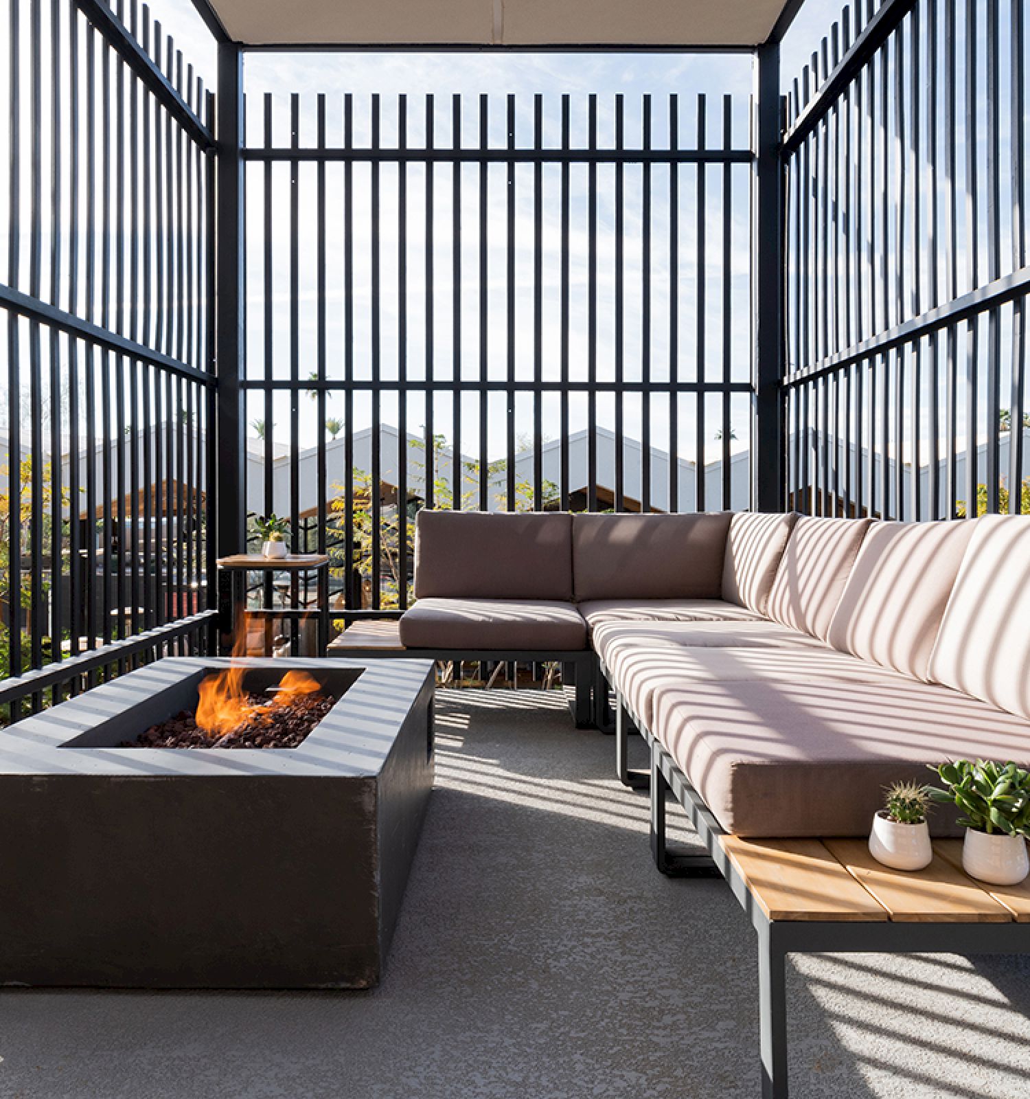 A modern outdoor seating area with a cushioned L-shaped couch, a fire pit in the center, and two small potted plants on a side table.