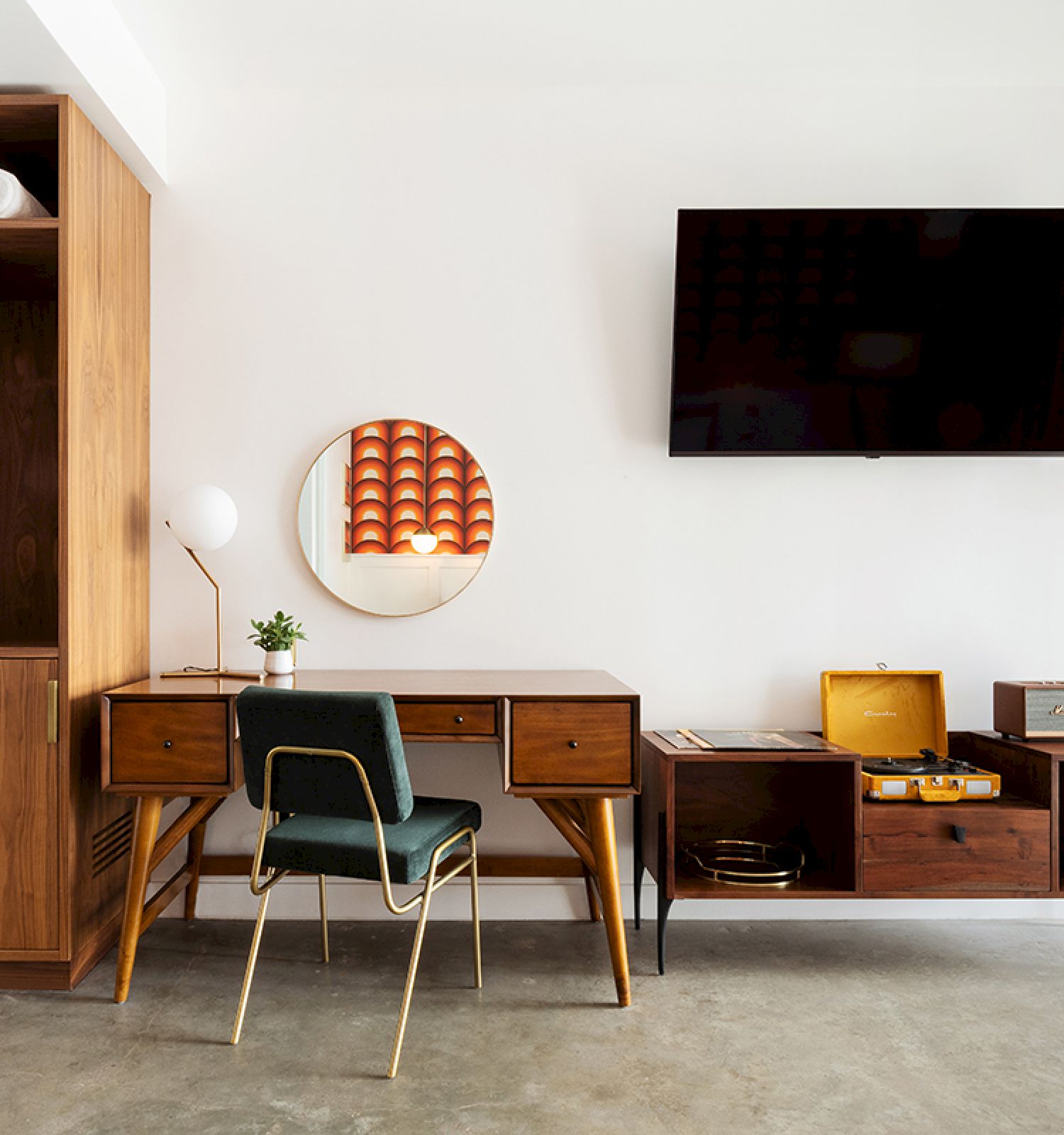 A modern room features wooden furniture, a TV on the wall, a desk with a chair, shelves with clothes and towels, and a hanging plant.