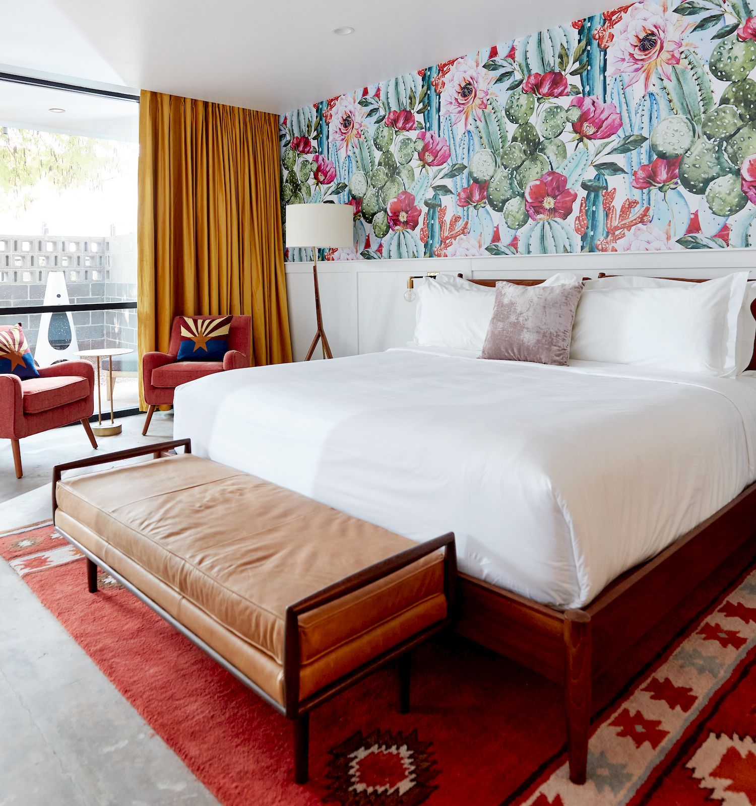 A stylish bedroom with a cactus-themed wallpaper, white bed, wooden furniture, a red rug, and large windows leading to an outdoor seating area.