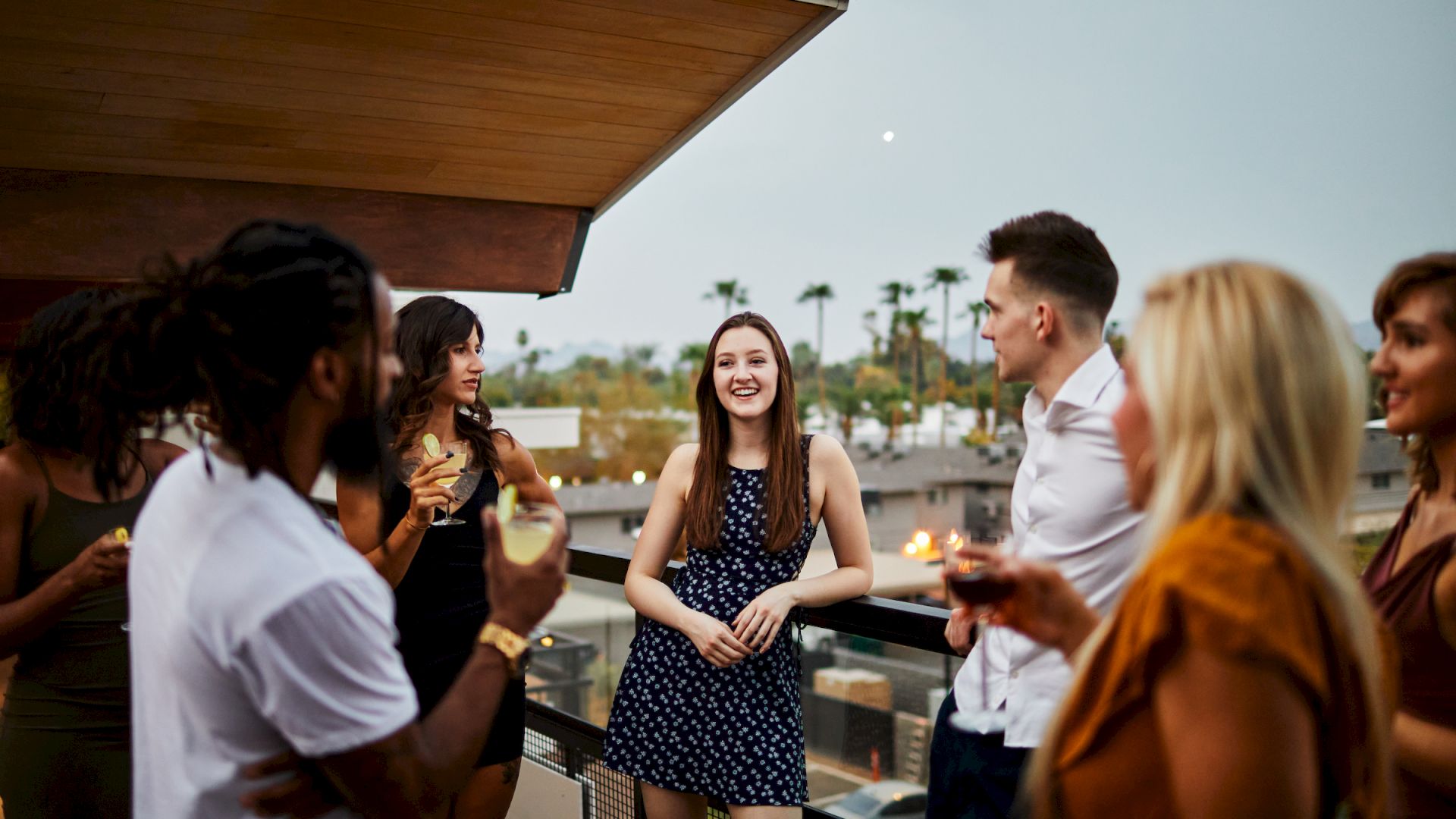 A group of people socializing on a rooftop, with some holding drinks, while the background shows palm trees and a twilight sky.