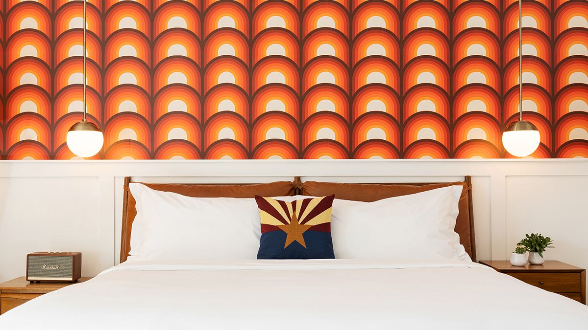 A neatly made bed with white linens, a decorative pillow, and two pendant lights in front of a retro, orange patterned wallpaper.