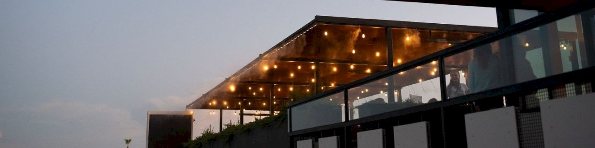 A rooftop patio with string lights, surrounded by glass railings and set against a twilight sky.