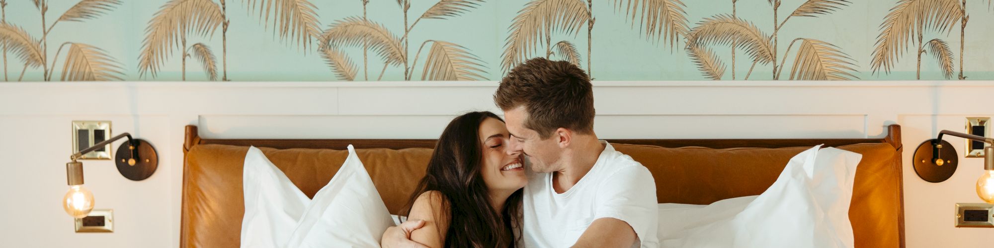 A couple sits close on a bed, kissing and holding hands; tropical palm wallpaper behind, cozy hotel vibes.