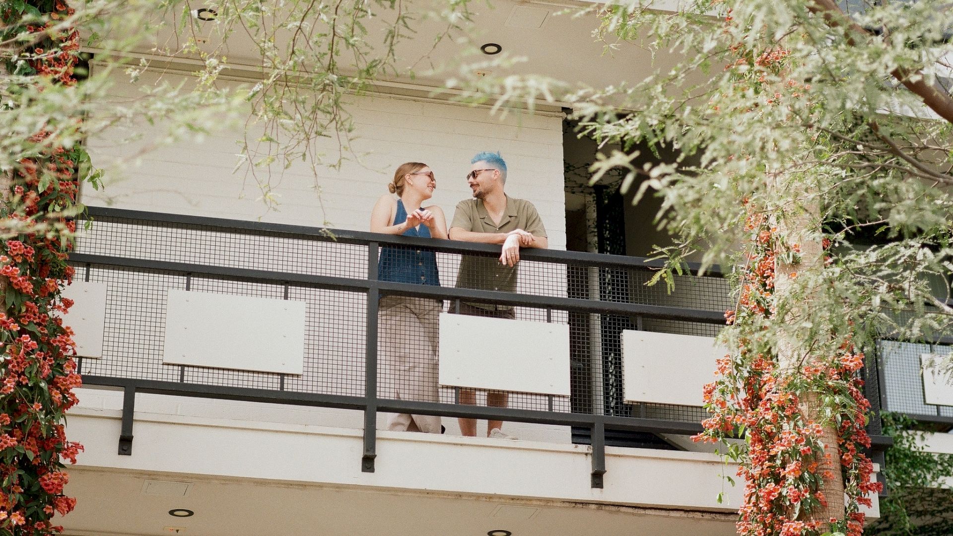 Two people are talking on a balcony surrounded by trees and plants, attached to a modern building.