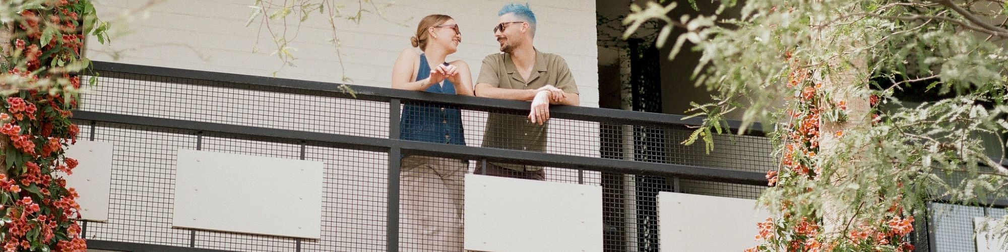 Two people are talking on a balcony surrounded by trees and plants, attached to a modern building.