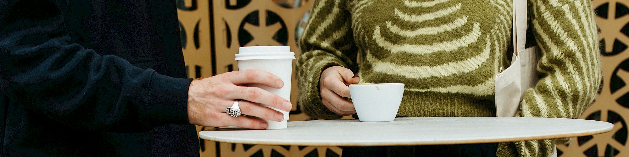 Two people standing at a round table outdoors, one holding a coffee cup, wearing casual clothing and sneakers, background with decorative pattern.