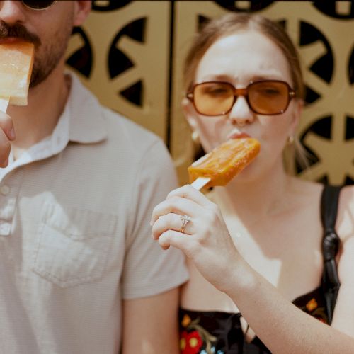 Two people wearing sunglasses enjoy popsicles in front of a patterned background, with the sun casting shadows on them.