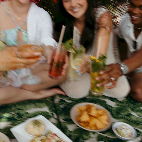 A group of people enjoying drinks and snacks outdoors, sharing a joyful moment together on a leafy-patterned blanket.