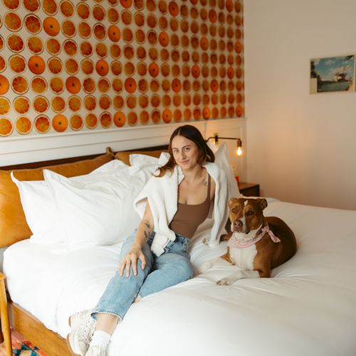 A person and a dog sit on a bed with white linens, against a wall decorated with vibrant orange slices.