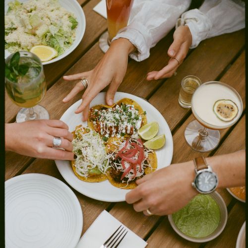 A table scene with tacos, lime wedges, cocktails, salad, and dips. Hands are reaching for food, creating a lively dining atmosphere.