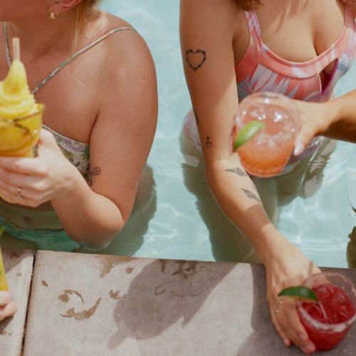 People enjoying colorful drinks while relaxing in a pool, with visible tattoos and swimwear, captured from the shoulders down.