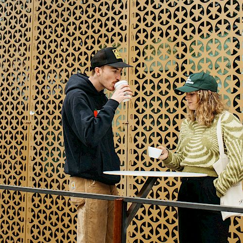 Two people are standing by a table and drinking from cups in front of a decorative lattice wall, both wearing hats.