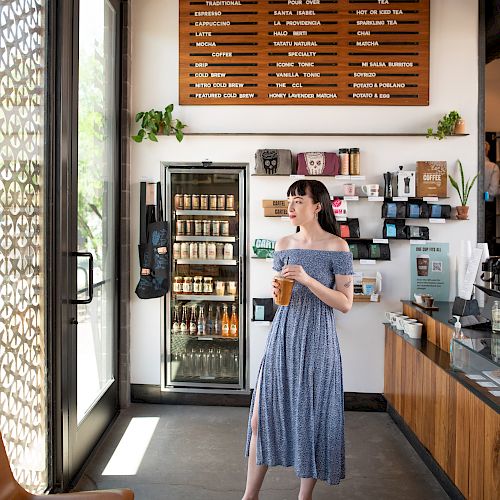 A woman stands in a coffee shop holding a drink. The menu board is visible behind her, along with shelves and a refrigerated display.