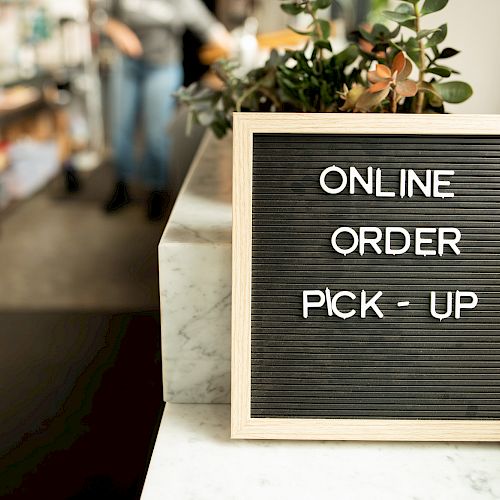 The image shows a sign on a counter that reads "ONLINE ORDER PICK - UP" with a plant nearby and people blurred in the background.