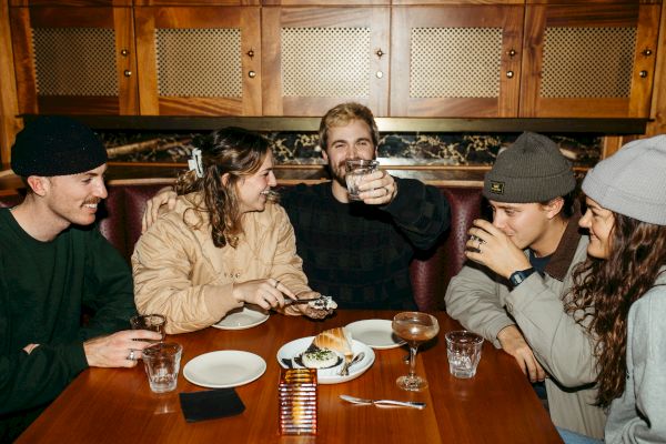 Five people are sitting together at a table, enjoying drinks, food, and sharing a laugh in a cozy setting.