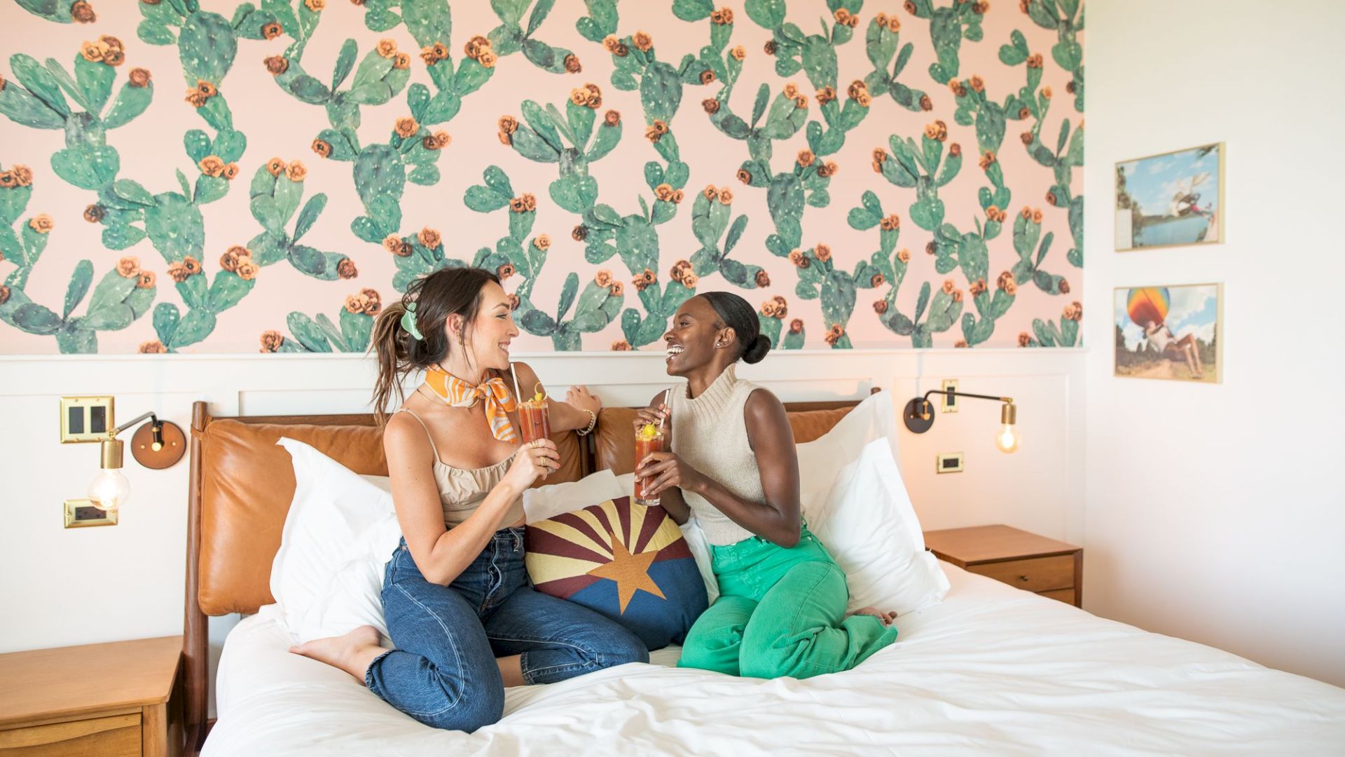 Two women are sitting on a bed, enjoying popsicles, with a cactus-themed wallpaper and colorful room decor in the background.