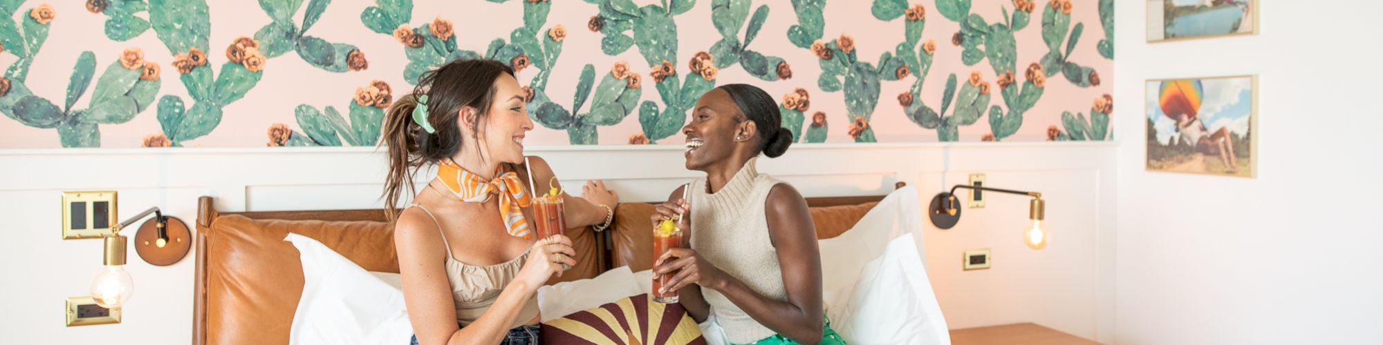 Two women are sitting on a bed, enjoying popsicles, with a cactus-themed wallpaper and colorful room decor in the background.