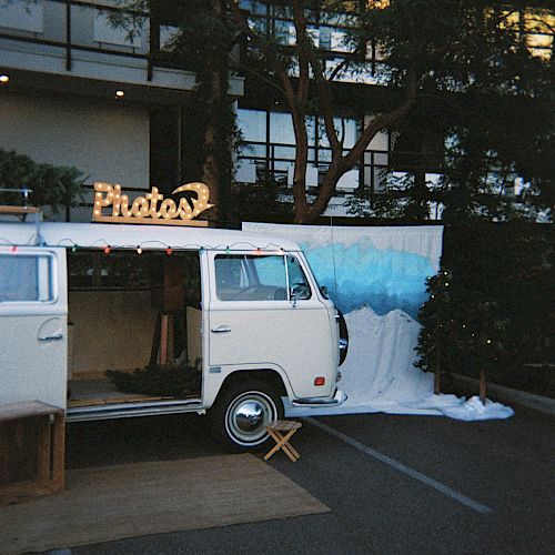 A vintage van converted into a photo booth with a snowy backdrop, parked near a building and surrounded by trees.