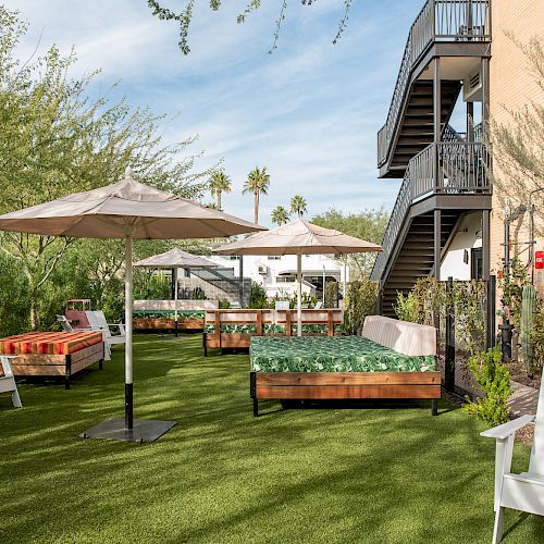 A grassy outdoor area with beds, umbrellas, and chairs, surrounded by trees and a staircase on the side of a building.