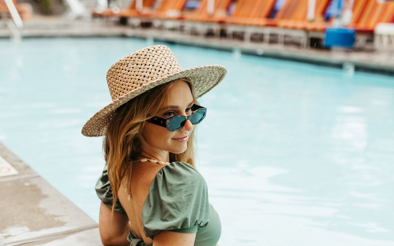 A person in a straw hat and sunglasses is by a pool, with orange loungers in the background.