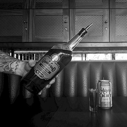 This image shows a person holding a bottle of Gran Centenario tequila over a table with a shot glass and a can of Tecate beer next to it.