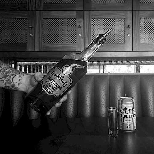 This image shows a person holding a bottle of Gran Centenario tequila over a table with a shot glass and a can of Tecate beer next to it.