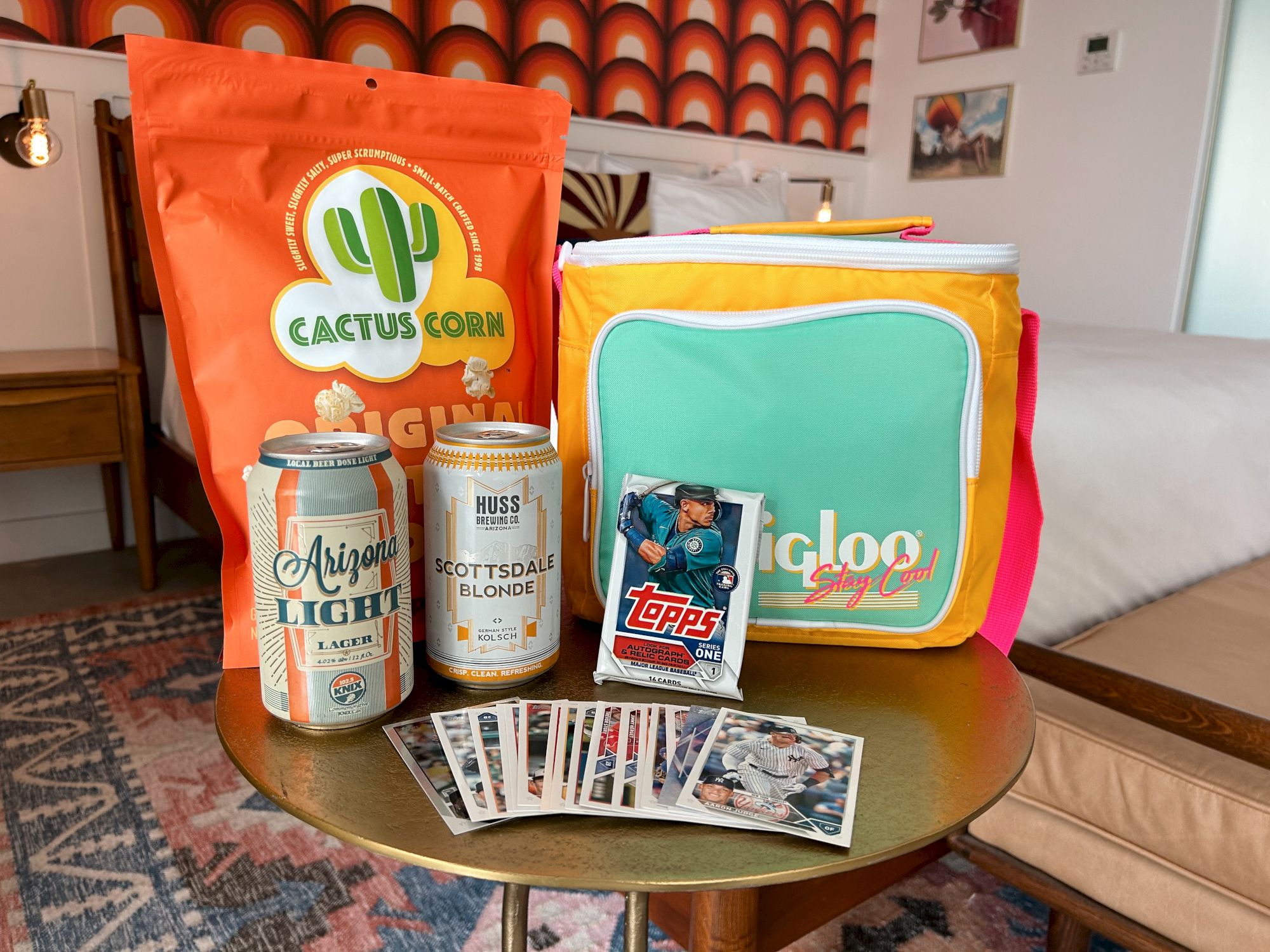 Snacks, drinks, sports cards, and a colorful cooler bag are displayed on a small table in a cozy room.
