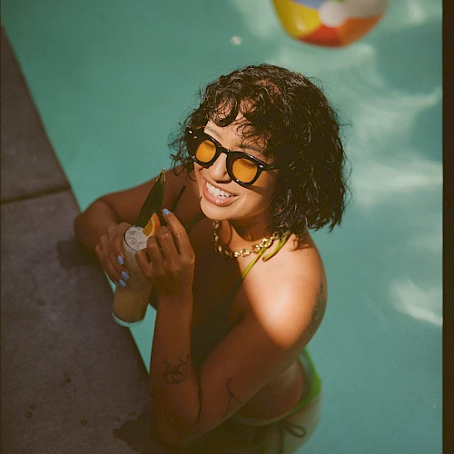 A woman with curly hair wearing sunglasses sits poolside in a bikini, smiling while giving a thumbs-up as a colorful beach ball floats nearby.