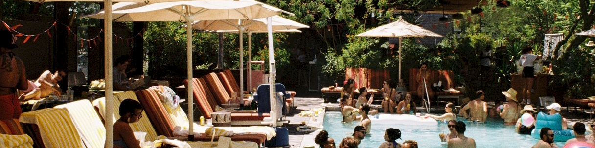 People lounge under shade sails and umbrellas by a sunny pool, a lively scene of vacation vibes and bright turquoise water.