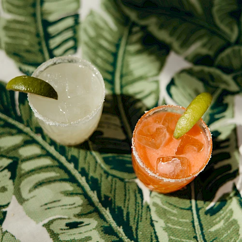 Two cocktails with lime wedges on a leafy patterned surface, one clear and one orange.
