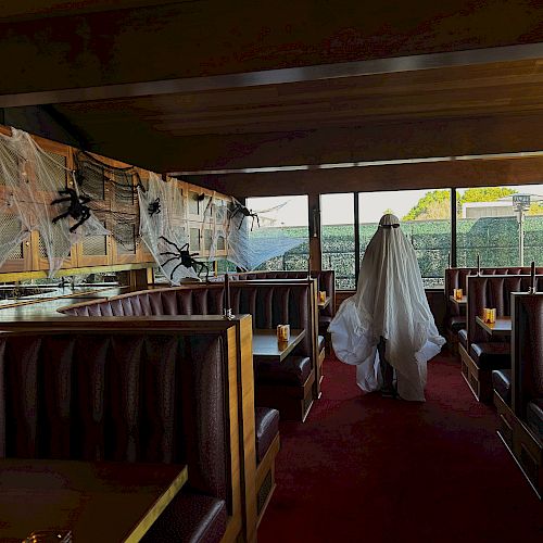 A person in a ghost costume stands inside a restaurant decorated with cobwebs, next to large windows providing a view.