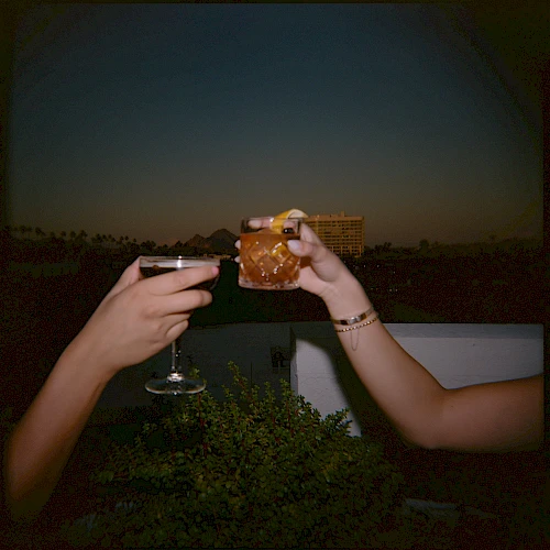 Two people clinking drinks on a balcony at dusk, toasting with a wine glass and a whiskey/tea glass, outdoors with city lights.