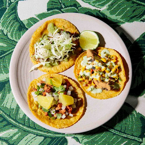 A plate with three tacos, each with different toppings, and a lime wedge sits on a leaf-patterned tablecloth.