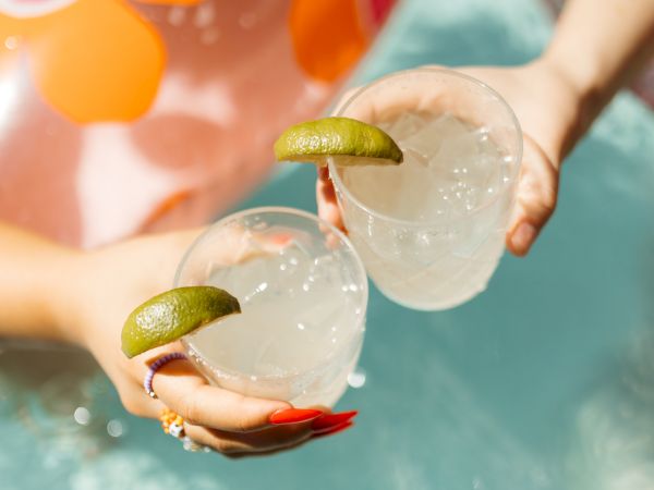 Two hands holding drinks garnished with lime slices over a pool, with an inflatable in the background.