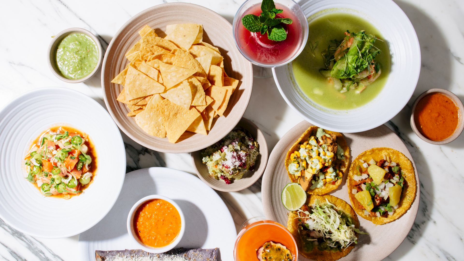 The image shows a variety of dishes including nachos, tacos, soup, a quesadilla, and several sauces on a marble surface.