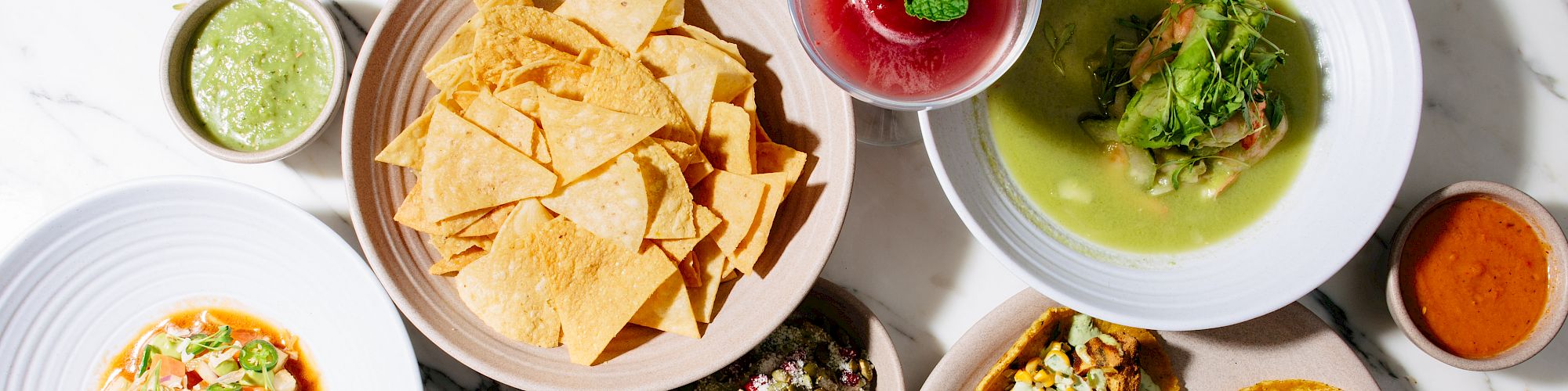 The image shows a variety of dishes including nachos, tacos, soup, a quesadilla, and several sauces on a marble surface.