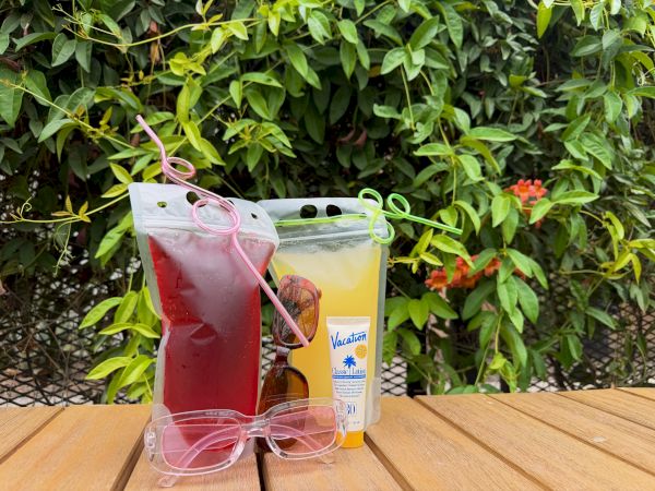 Two juice pouches (red and yellow) with a straw, a pair of pink glasses, and a small bottle on a wooden table outdoors.