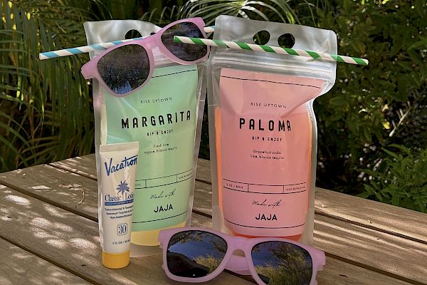 Two drink pouches, sunscreen, and two pairs of sunglasses sit on a wooden surface, surrounded by greenery and sunlight.