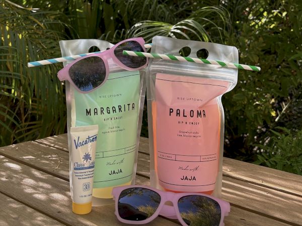 Two drink pouches, sunscreen, and two pairs of sunglasses sit on a wooden surface, surrounded by greenery and sunlight.