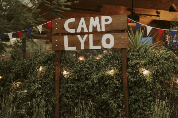 A rustic wooden sign with "CAMP LYLO" written on it, surrounded by greenery and festive string lights, creates a cozy outdoor atmosphere.